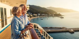 A senior couple in their late sixties standing on the deck of a cruise ship near Las Vegas, enjoying the best cruise lines for seniors experience as they depart from Southern California with warm afternoon sunlight over the coastal harbor.