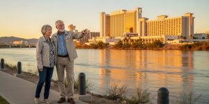 A senior couple enjoying a relaxed walk along the flat paved Riverwalk beside the Colorado River in Laughlin Nevada, with casino hotels reflected in the calm water behind them — the kind of peaceful seniors overnight getaway that Las Vegas locals have discovered just 97 miles from the Strip.